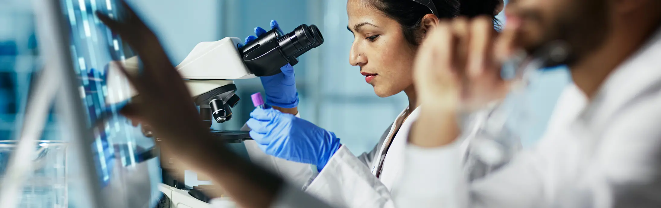 Un laboratorio con una mujer sentada delante de un microscopio mirando una muestra, a su lado, un hombro con barba mira una pantalla