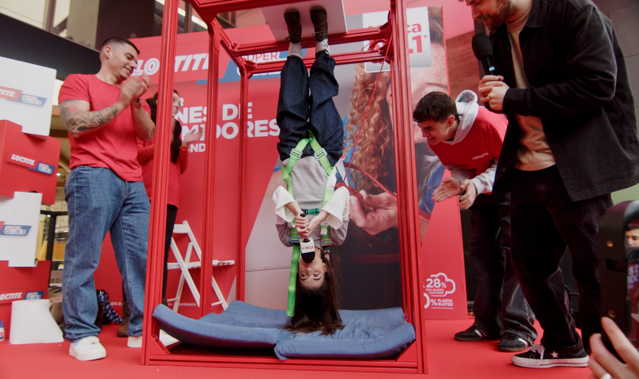 Persona sujeta boca abajo en la estructura de la demostración “Hanging Man” de Loctite SuperGlue‑3, rodeada por equipo y asistentes en el evento de L’Illa Diagonal.