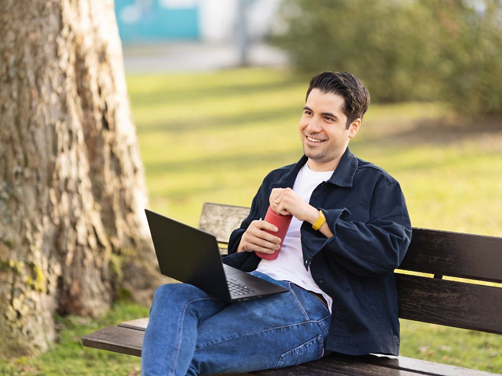 Un empleado de Henkel se sienta en un banco de un parque y trabaja con su portátil.