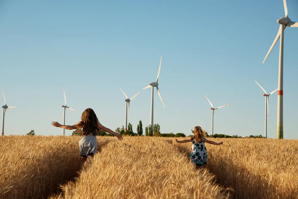 Unas niñas corren por un campo hacia unas turbinas eólicas.