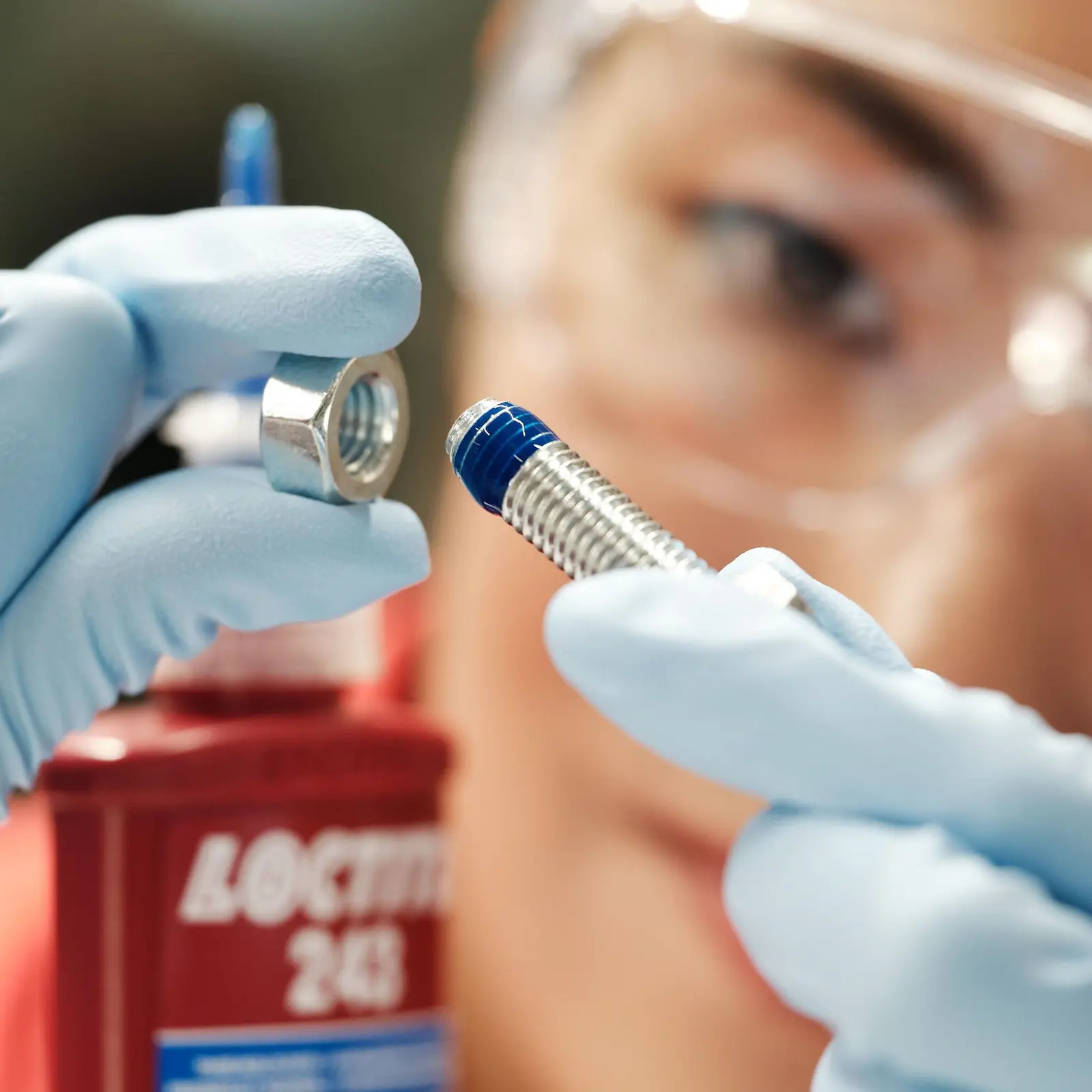 En el fondo, el rostro de una mujer. Al frente, sus manos con guantes azules sosteniendo un tornillo con Threadlocker azul.