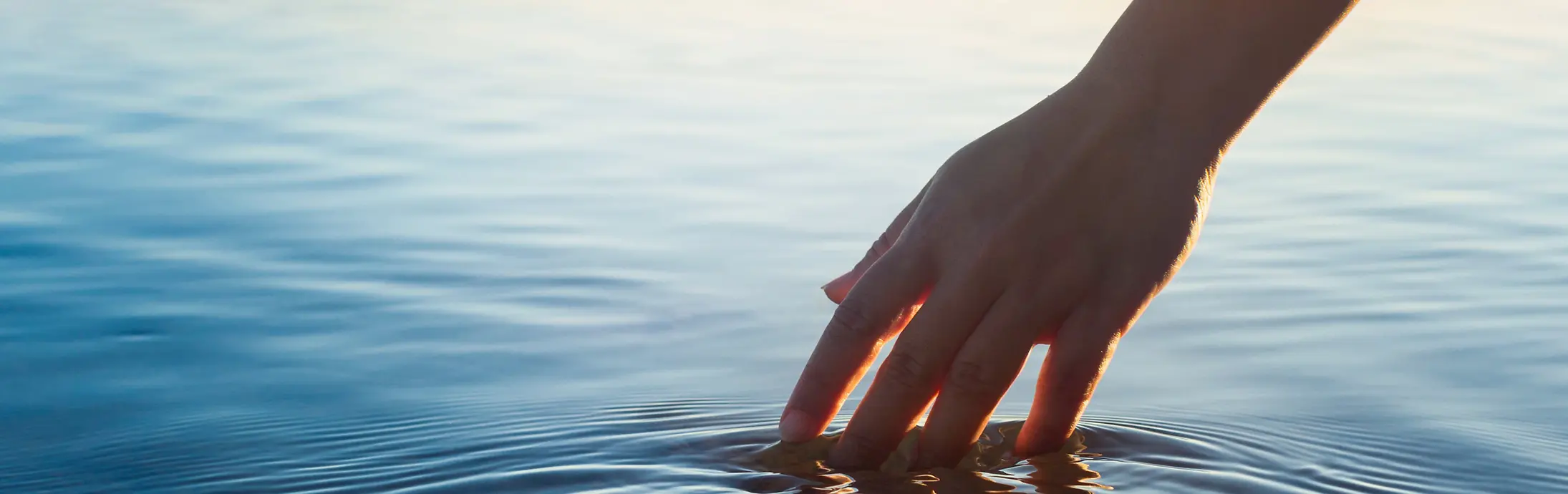 Una mano acaricia una superficie de agua en calma frente al horizonte