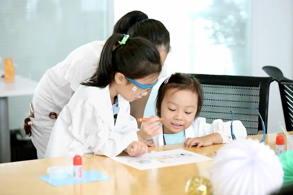 Dos niñas y una mujer con bata científica colorean en una mesa