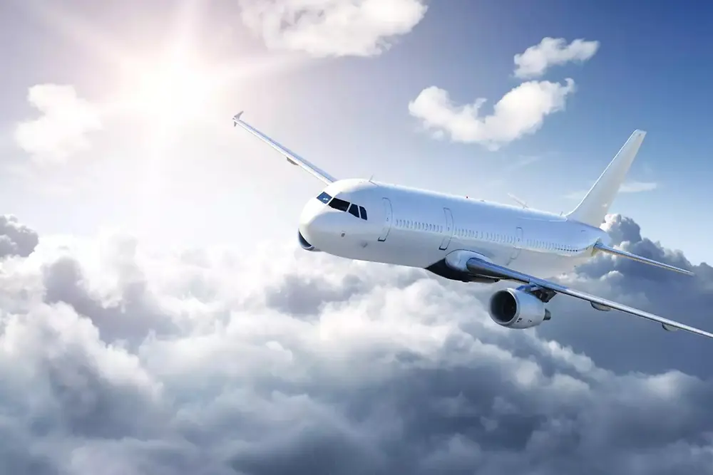 Un avión de pasajeros blanco vuela sobre nubes suaves en un cielo brillante, con la luz del sol desde la parte superior izquierda.