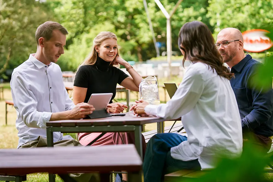 Grupo de compañeros sentados juntos al aire libre, hablando del trabajo con tabletas y ordenadores portátiles, lo que representa la colaboración y una cultura abierta y orientada al equipo en Henkel.