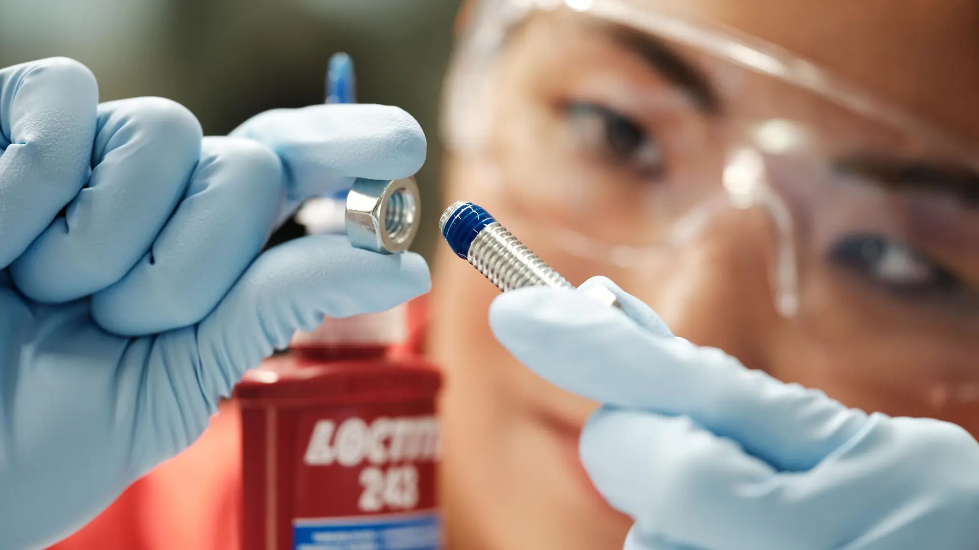 En el fondo, el rostro de una mujer. Al frente, sus manos con guantes azules sosteniendo un tornillo con Threadlocker azul.