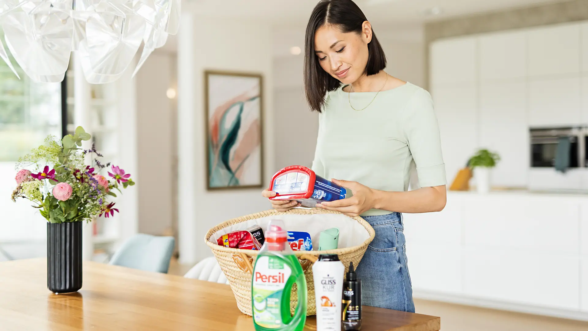 Mujer con una cesta de pie sobre una mesa que está llena de productos Henkel como: Persil, Gliss, Bref, Somat, taft y más.