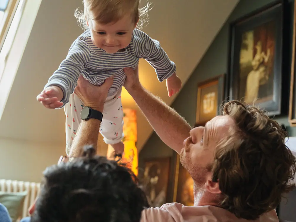 Dos padres sostienen a su bebé y sonríen en casa, lo que simboliza la inclusión y la política de permiso parental de Henkel, favorable para las familias.
