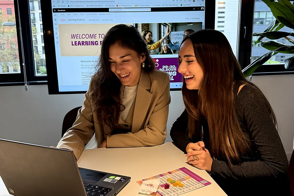 Dos empleadas frente a un portátil con folletos del Learning Festival, pantalla al fondo mostrando la página del evento.