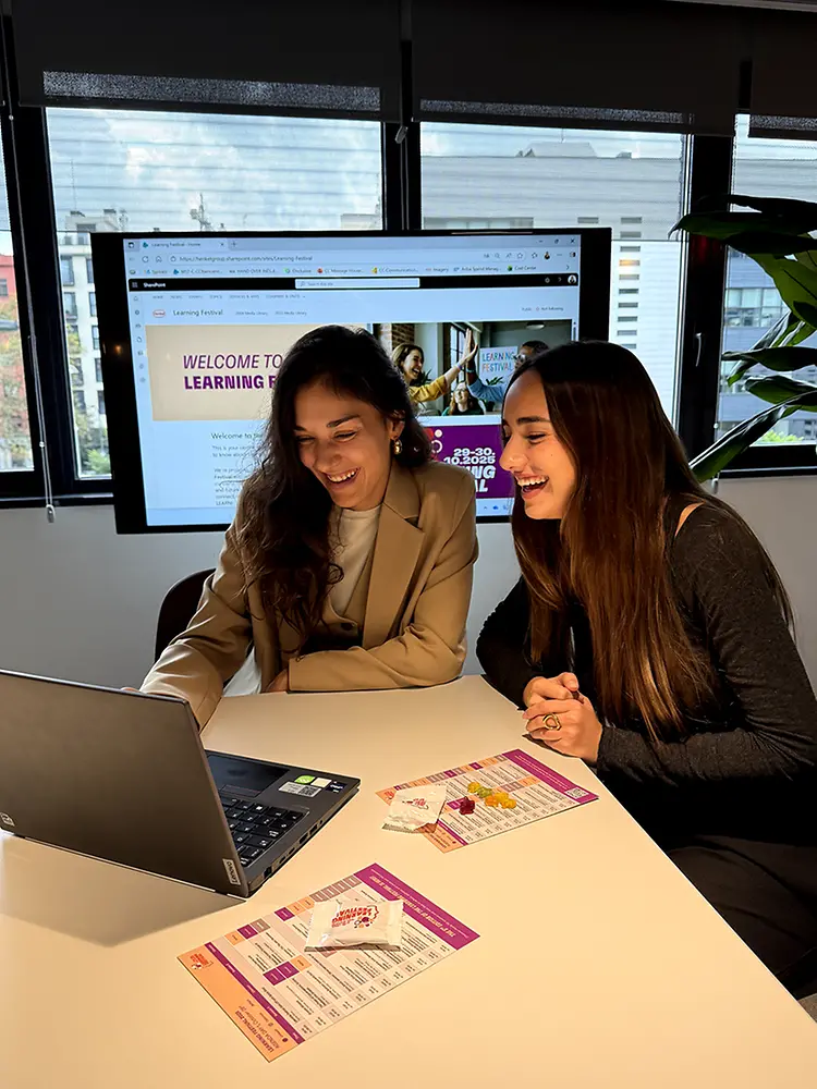Dos empleadas frente a un portátil con folletos del Learning Festival, pantalla al fondo mostrando la página del evento.