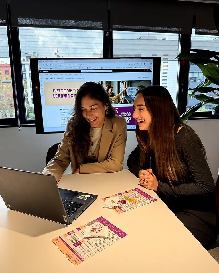Dos empleadas frente a un portátil con folletos del Learning Festival, pantalla al fondo mostrando la página del evento.