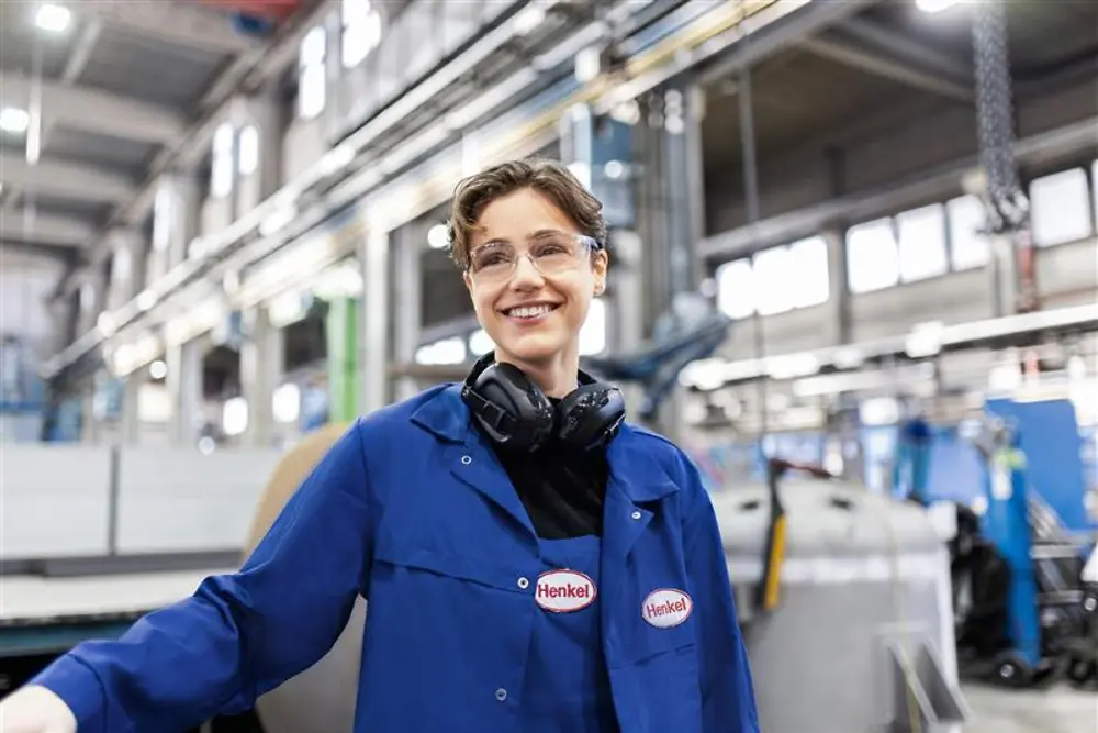 Empleado sonriente de Henkel con una bata de trabajo azul y auriculares de seguridad, de pie en una nave de producción industrial, lo que representa la experiencia y la innovación en la fabricación.