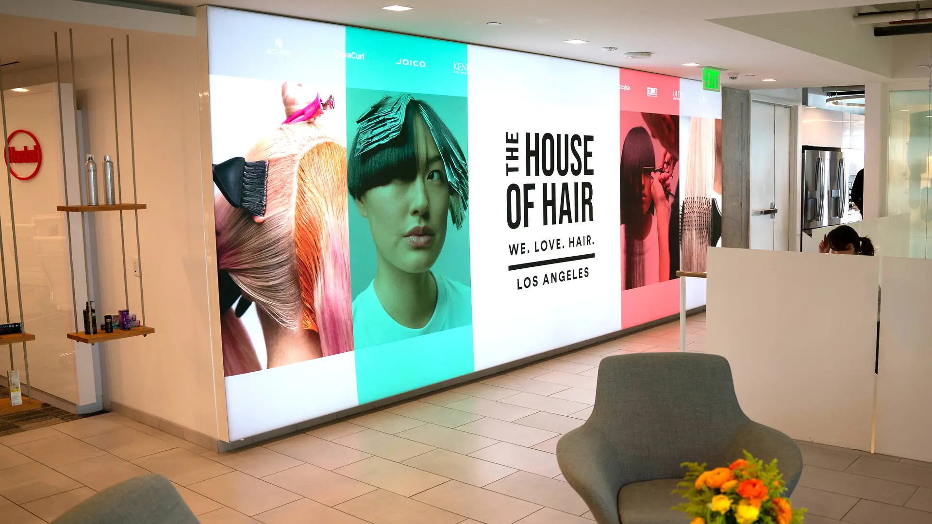 Un vestíbulo moderno con una pared luminosa que muestra grandes paneles con gráficos coloridos inspirados en el cabello y el texto “THE HOUSE OF HAIR – WE. LOVE. HAIR. – LOS ANGELES.” El espacio incluye sillones de color gris claro, una mesa pequeña con un arreglo floral vibrante y un diseño limpio y minimalista con suelo embaldosado y paneles blancos.