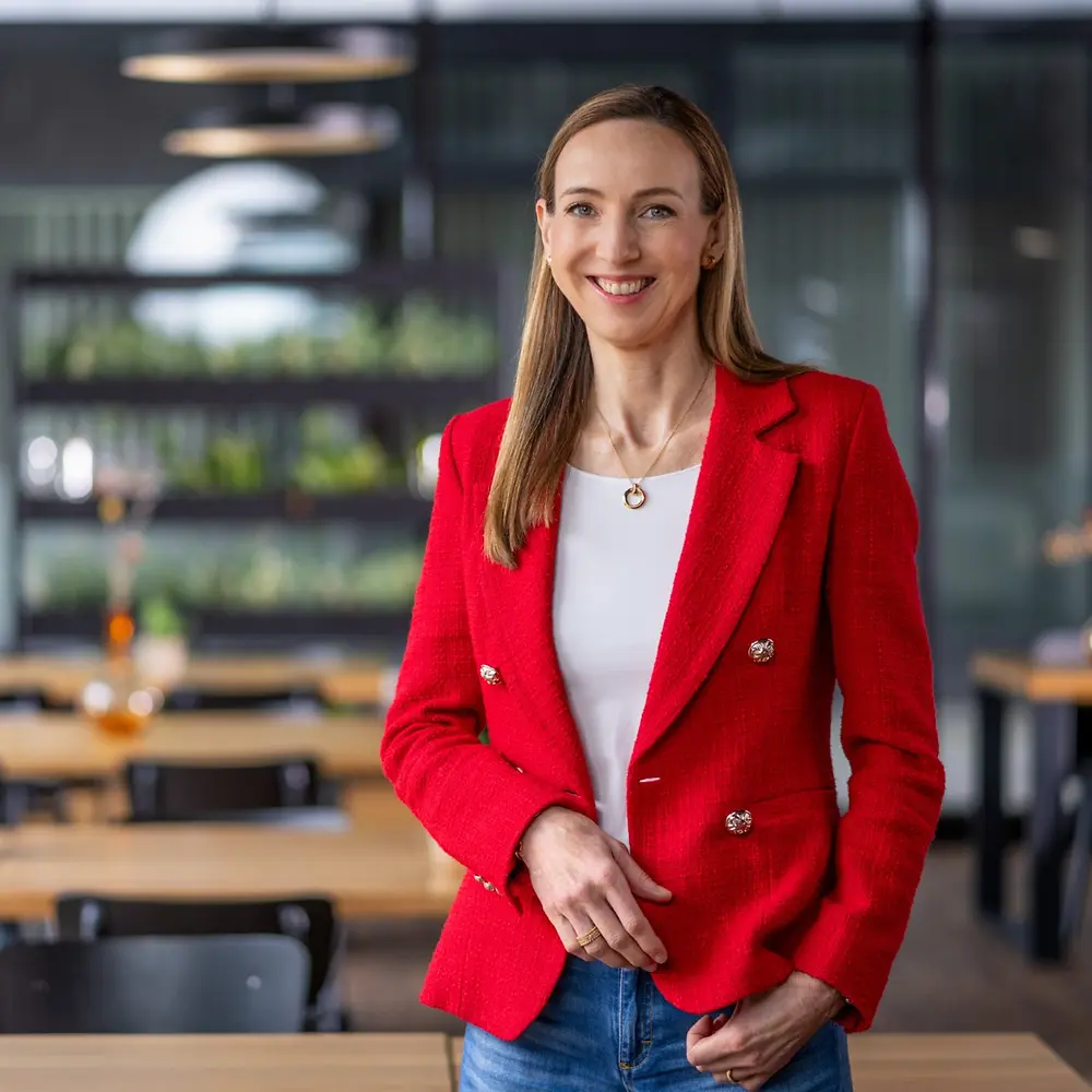 Dr. Simone Bagel-Trah con blazer rojo brillante y blusa blanca, de pie en un entorno moderno estilo café con mesas de madera y jarrones decorativos al fondo.