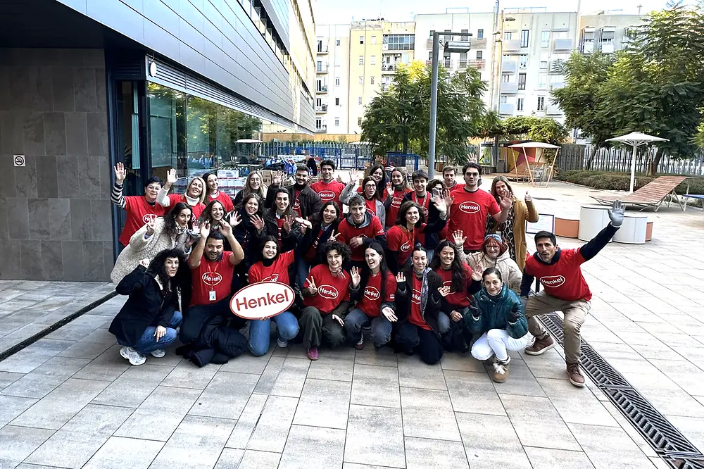 Grupo de empleados de Henkel Ibérica con camisetas rojas reunidos en el exterior de la sede en Barcelona, sosteniendo un cartel con el logotipo de Henkel, durante el mercadillo solidario anual.