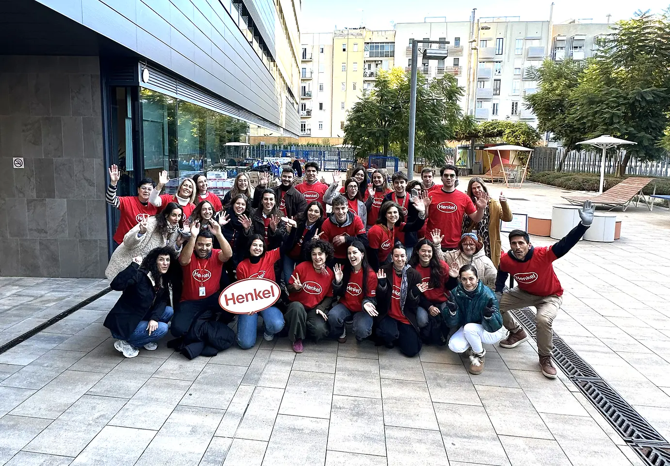 Grupo de empleados de Henkel Ibérica con camisetas rojas reunidos en el exterior de la sede en Barcelona, sosteniendo un cartel con el logotipo de Henkel, durante el mercadillo solidario anual.