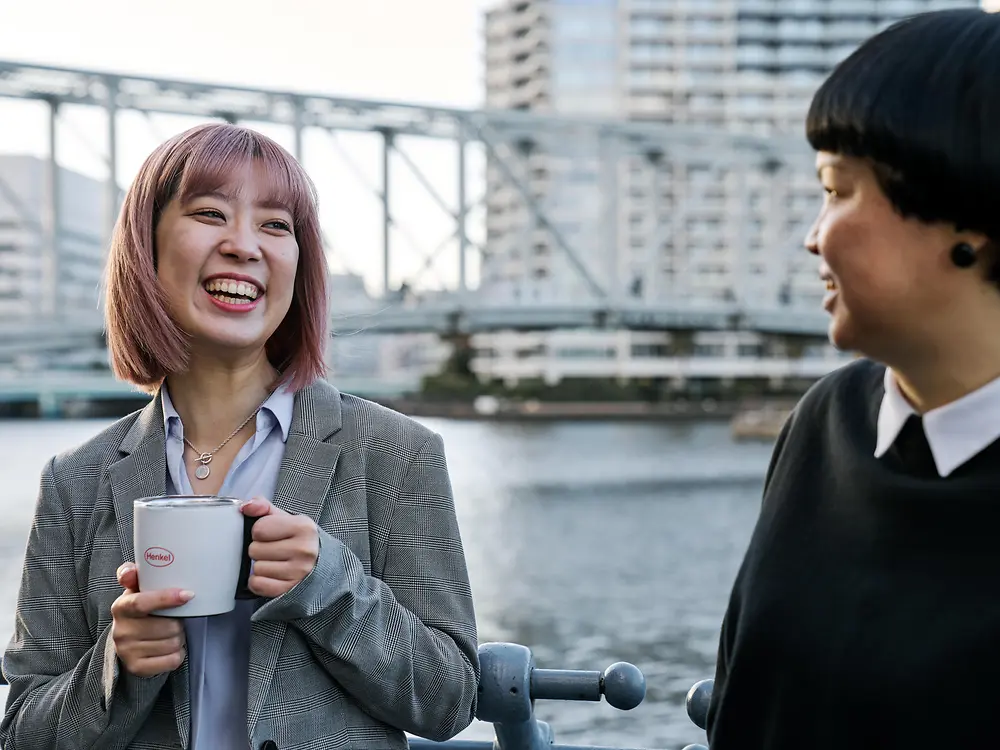Dos personas mantienen una conversación informal junto a una barandilla frente al mar, una de ellas sostiene una taza de café con la marca Henkel, con un puente y edificios de la ciudad al fondo, lo que ilustra la cultura de trabajo flexible de Henkel.