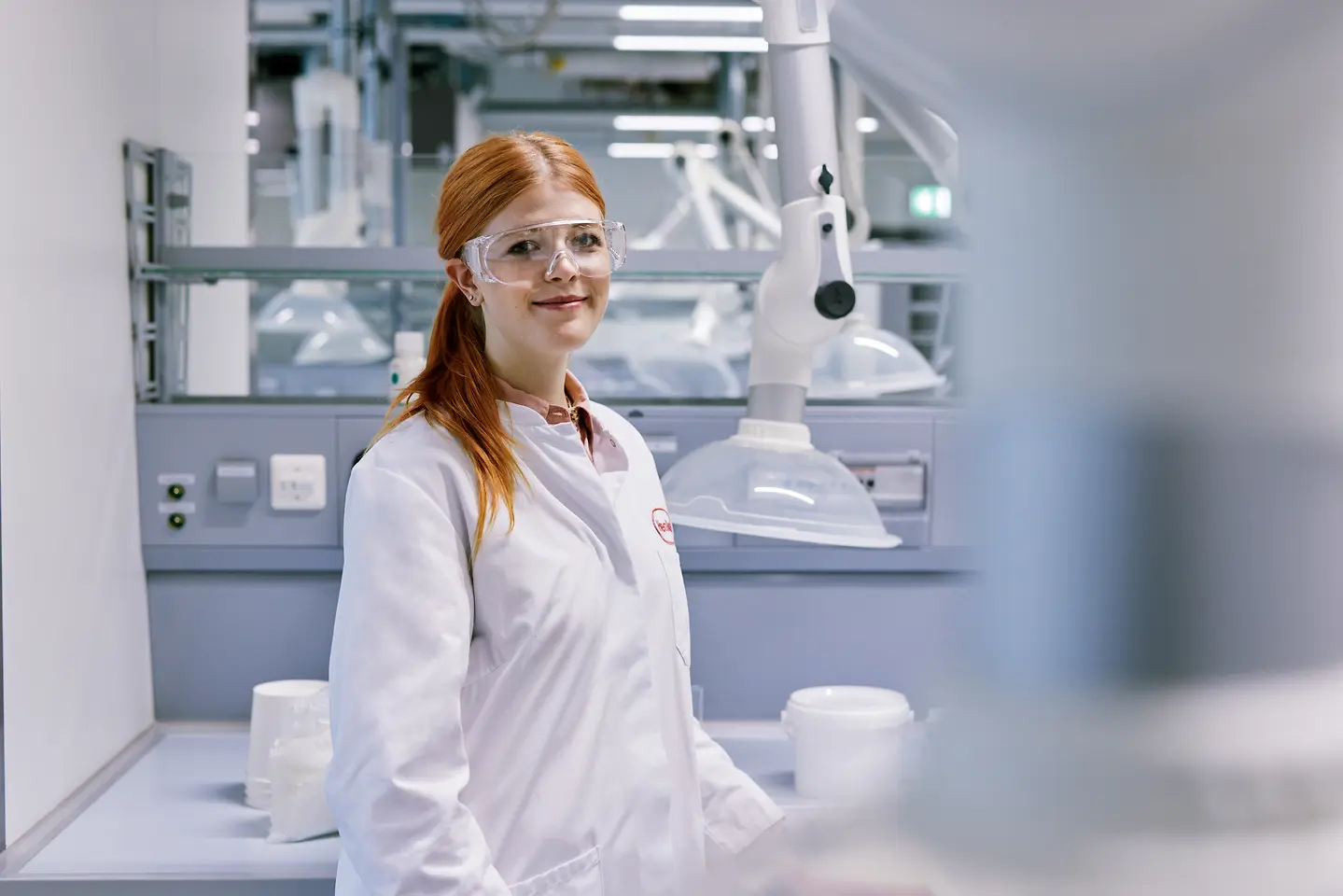 Graduado que comienza su carrera en Henkel, con una bata de laboratorio blanca y de pie en un moderno entorno de laboratorio con equipos técnicos al fondo.