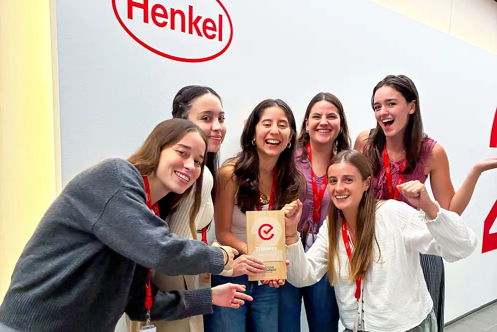 Un grupo de seis personas está de pie frente a una pared con el logotipo de Henkel. Llevan cordones rojos y ropa informal, y están muy juntos alrededor de una placa de premio. El entorno parece ser un espacio corporativo interior con una pared de color claro y suelo gris. El grupo posa con entusiasmo, destacando el premio en el centro.