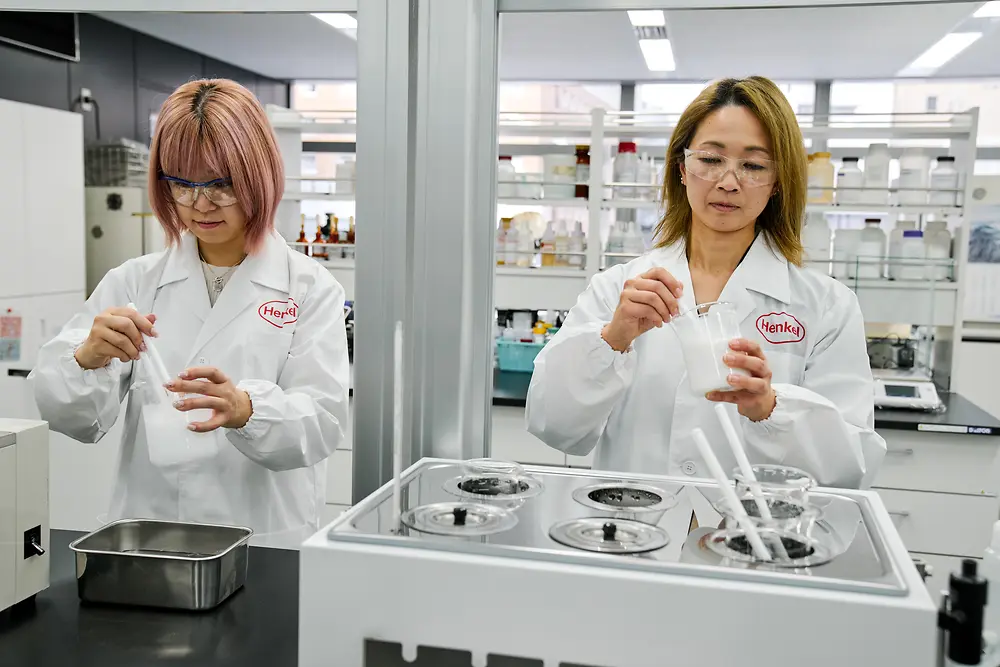 Dos personas con batas de laboratorio blancas de Henkel están trabajando en un laboratorio moderno. Están manipulando instrumentos científicos y recipientes sobre una encimera, con varios equipos de laboratorio y estanterías con suministros visibles al fondo.