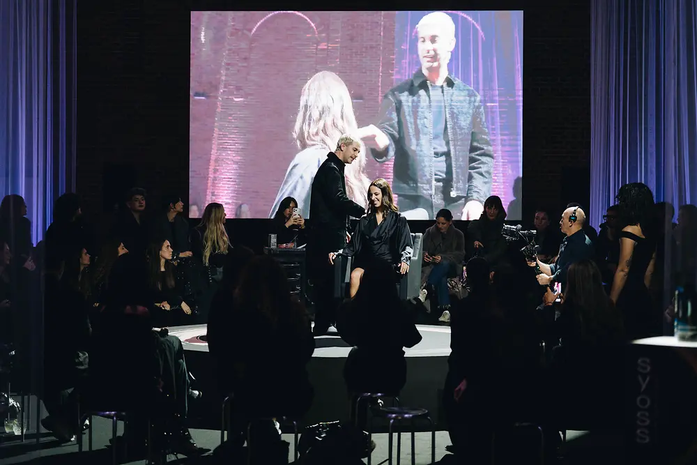 Evento de lanzamiento de Syoss en Madrid con una masterclass en directo. En la imagen se ve al estilista aplicando color sobre el escenario, rodeado de asistentes, con una gran pantalla proyectando la demostración.