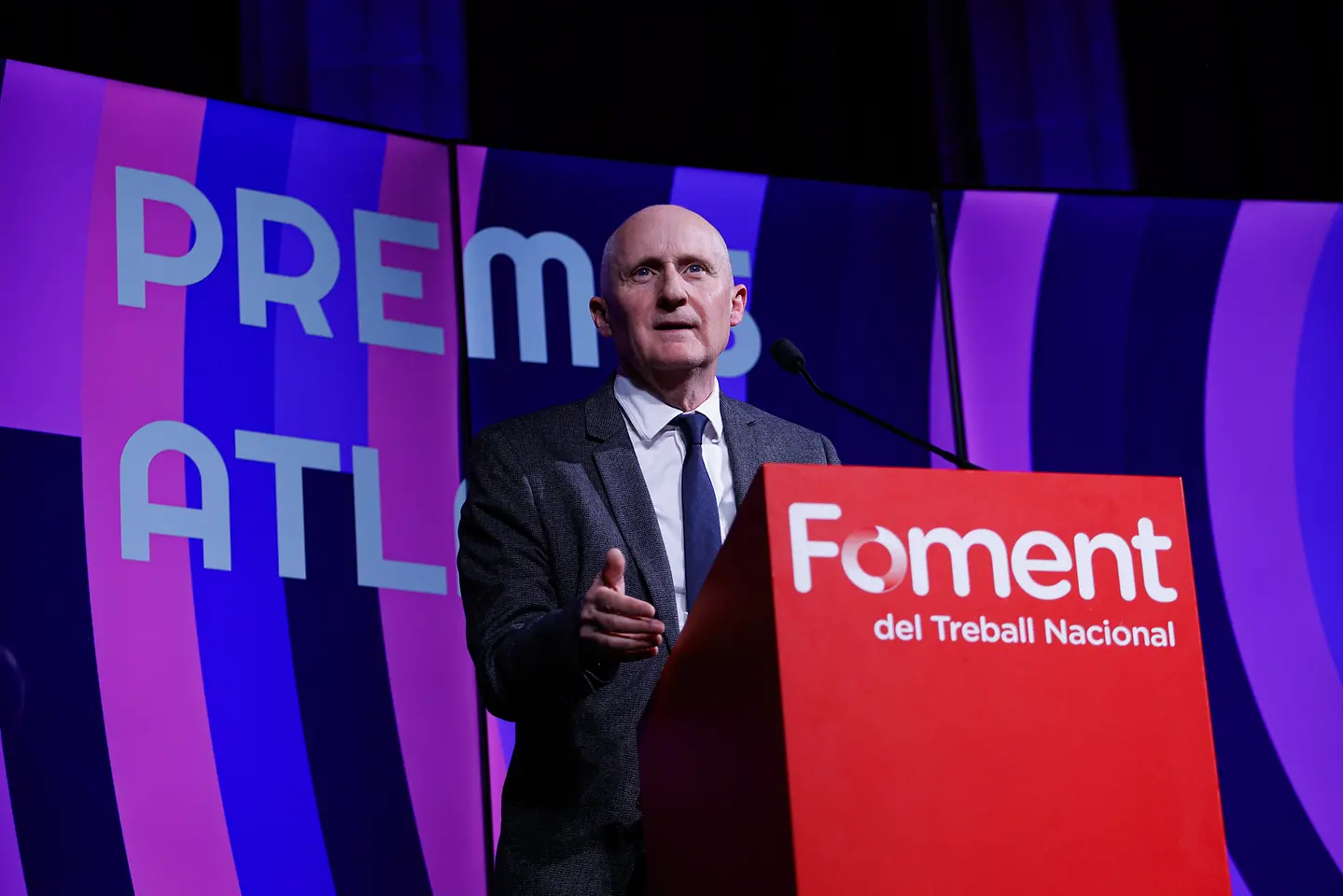 Miguel Rodríguez hablando en el escenario de los XII Premios Atlante, con un atril rojo que lleva el logotipo “Foment del Treball Nacional” y un fondo formado por paneles verticales en tonos morados y rosas con el texto “Premis Atlante”.