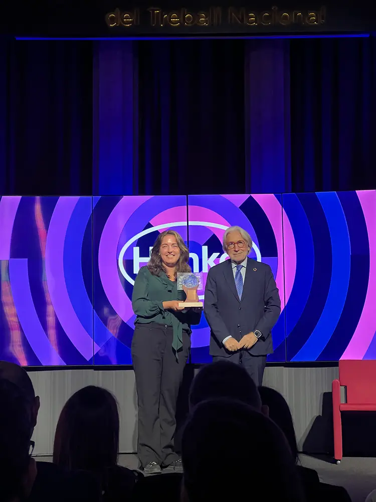Núria Vaqué sosteniendo el trofeo en el escenario de los XII Premios Atlante. Al fondo, una pantalla con diseño circular en tonos morados y rosas que muestra el logotipo de Henkel.