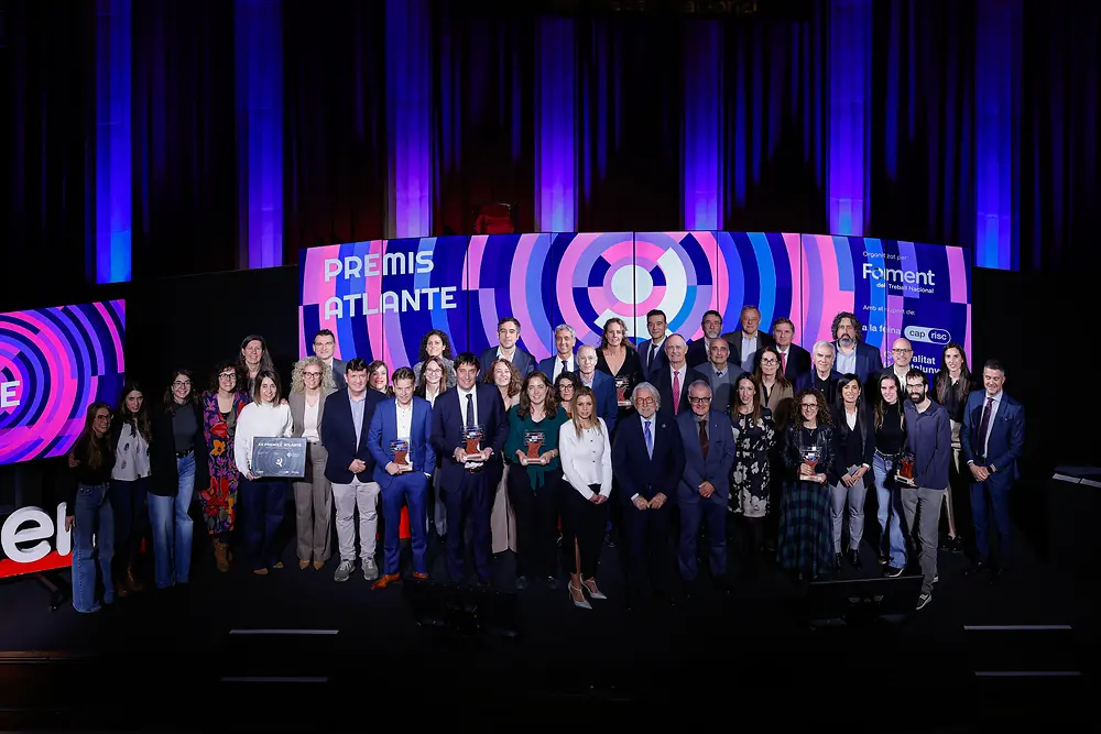Grupo de personas en el escenario de los XII Premios Atlante, con un fondo formado por pantallas en tonos morados y rosas que muestran los textos “Premis Atlante” y “Foment del Treball Nacional”. Varias personas sostienen trofeos en la parte frontal.