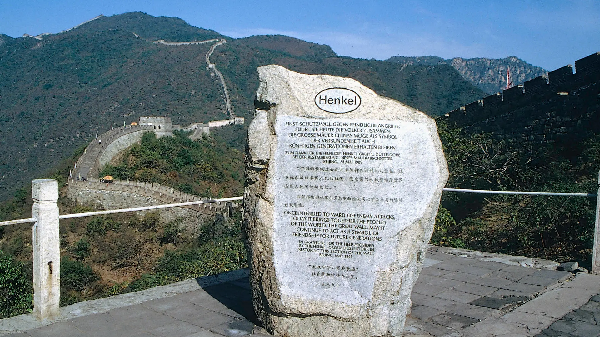 Una lápida conmemorativa que recuerda el apoyo de Henkel a la restauración de un tramo de la Gran Muralla China. Al fondo, la Gran Muralla China serpentea a través de una zona montañosa boscosa.