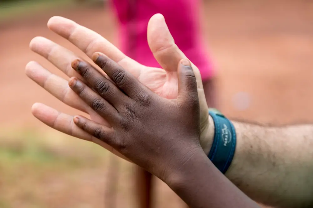 Primer plano de dos manos, una adulta y otra infantil, apoyadas entre sí en señal de unión y colaboración, sobre un fondo desenfocado al aire libre.