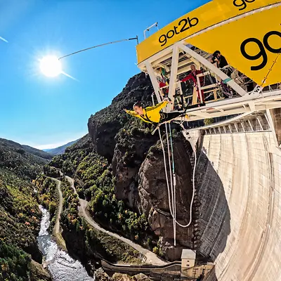 Influencer Laura Escanes realizando un salto de puenting desde una plataforma amarilla con el logo de got2b, sobre una presa rodeada de montañas y un río, en un día soleado.
