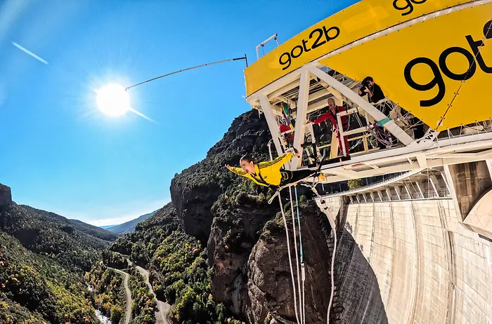 Influencer Laura Escanes realizando un salto de puenting desde una plataforma amarilla con el logo de got2b, sobre una presa rodeada de montañas y un río, en un día soleado.