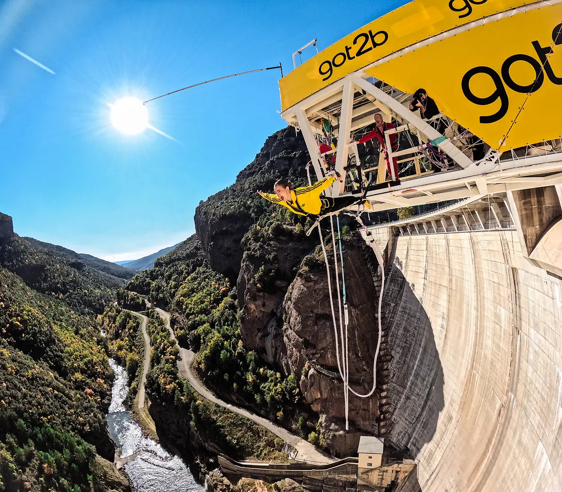 Influencer Laura Escanes realizando un salto de puenting desde una plataforma amarilla con el logo de got2b, sobre una presa rodeada de montañas y un río, en un día soleado.