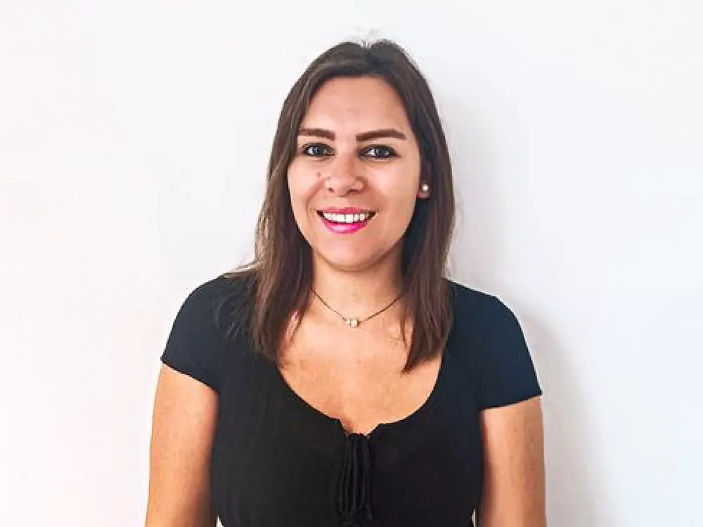Personal de selección de pie frente a un fondo blanco, con una camiseta negra y un cinturón beige, comparte consejos sobre cómo gestionar las preguntas difíciles de una entrevista.