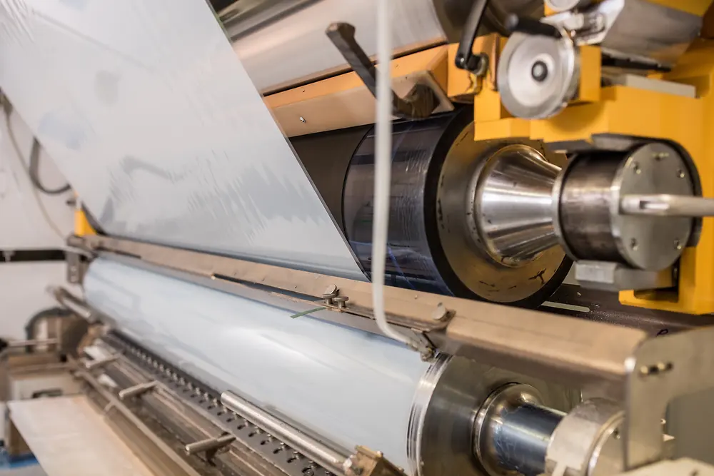 Máquina industrial aplicando o laminando una película adhesiva sobre un rodillo, con cilindros metálicos en funcionamiento en una línea de producción.