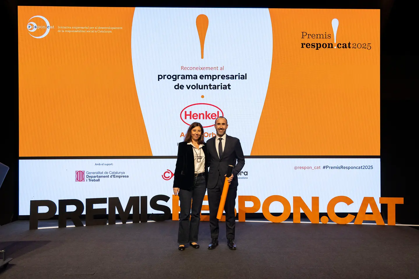 Dos personas posan en el escenario de los Premis Respon.cat 2025 frente a una pantalla que anuncia el reconocimiento a Henkel por su programa empresarial de voluntariado.