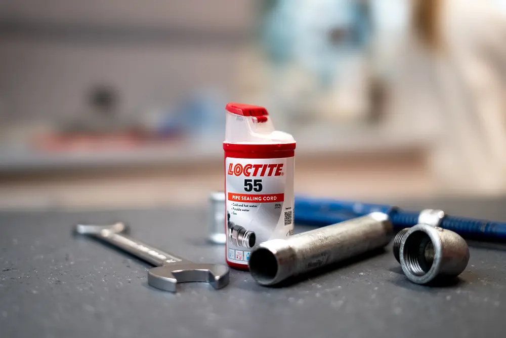 Envase de LOCTITE 55 sobre una superficie de trabajo junto a herramientas y piezas de tubería metálica, utilizado para el sellado de roscas en instalaciones de fontanería.