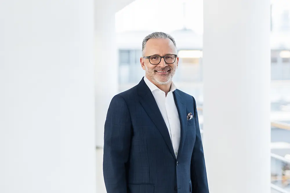 Retrato corporativo de Carsten Knobel, CEO de Henkel, sonriendo y vestido con traje azul oscuro y camisa blanca en un entorno de oficina luminoso.