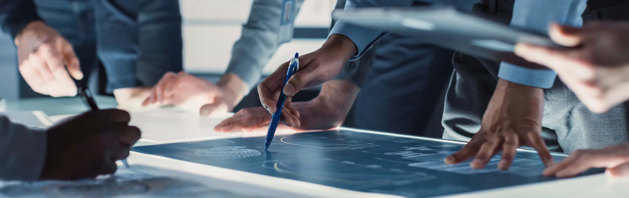 Vista en primer plano de varias personas reunidas alrededor de una gran mesa iluminada, revisando y marcando planos técnicos con bolígrafos y dispositivos digitales.