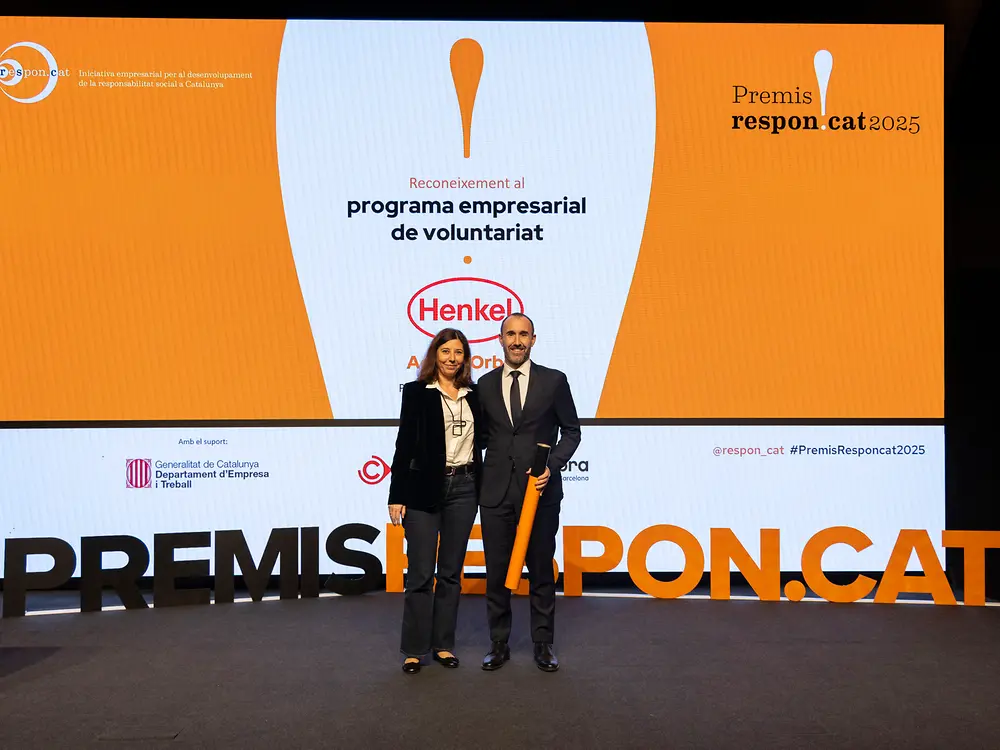 Imagen de dos personas sobre un escenario durante la ceremonia de los Premios Respon.cat, frente a una pantalla iluminada que muestra el logotipo de Henkel y la frase “programa empresarial de voluntariat”.