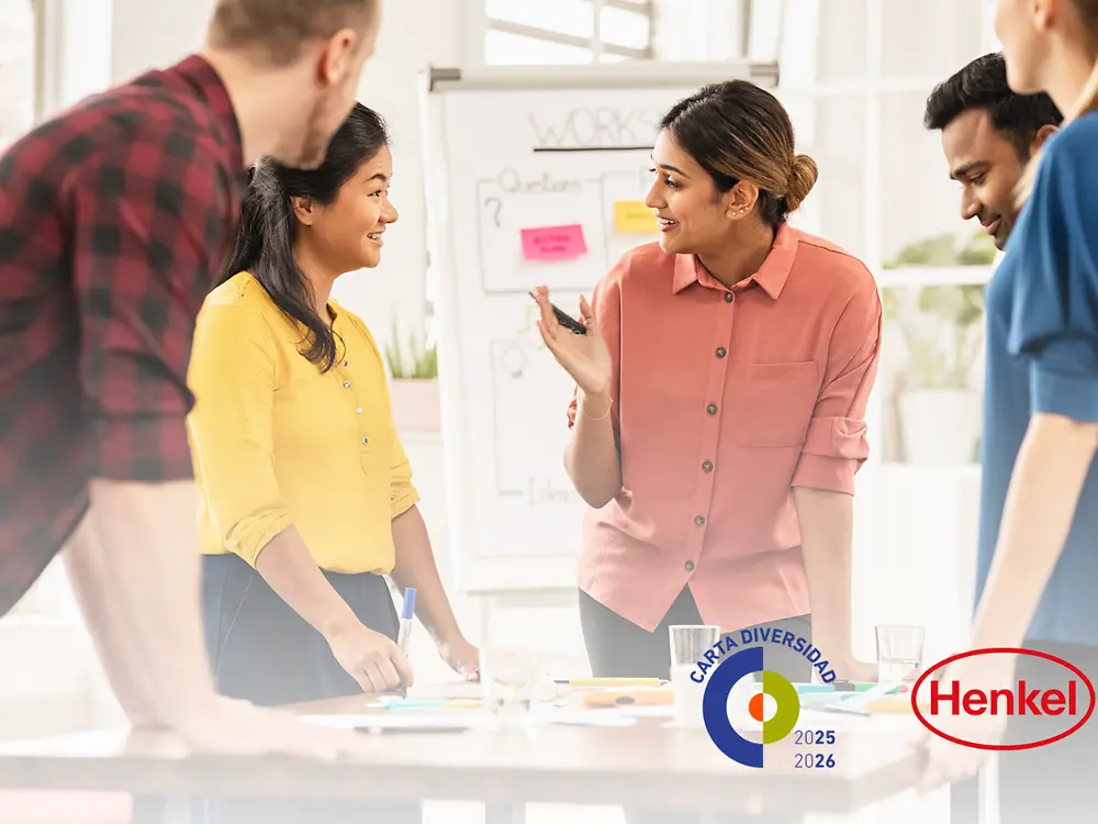 Imagen de varias personas reunidas alrededor de una mesa de trabajo, observando un tablero blanco con notas y materiales de apoyo, simbolizando el compromiso de Henkel Ibérica con la diversidad y la inclusión en el entorno laboral.