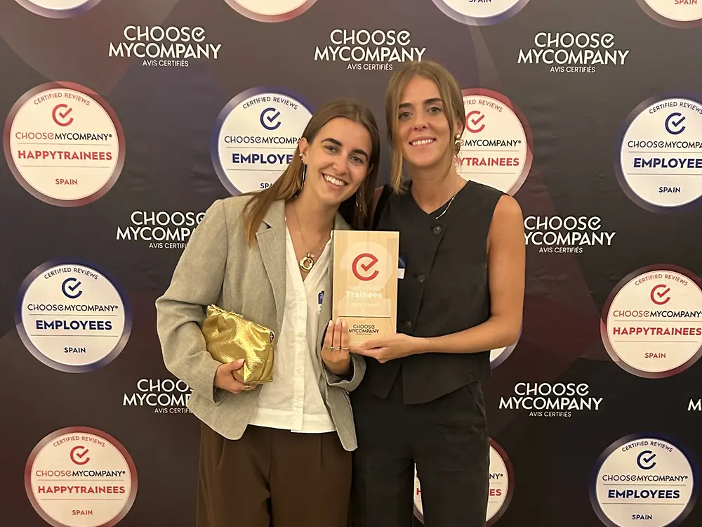 Imagen de dos empleadas sosteniendo un certificado HappyTrainees frente a un fondo decorado con varios logotipos relacionados con los premios, representando el reconocimiento otorgado a Henkel Ibérica por su labor en el desarrollo y bienestar de jóvenes talentos.