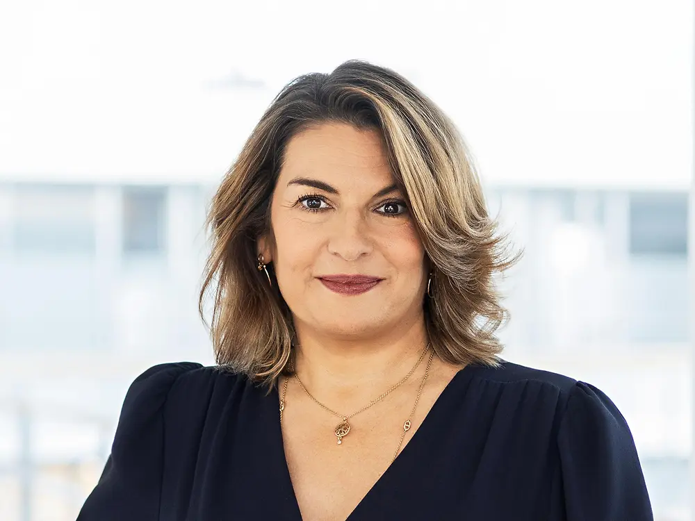 Retrato corporativo de Sylvie Nicol, directiva de Henkel, con blusa azul oscuro y cabello castaño, posando en una oficina luminosa.