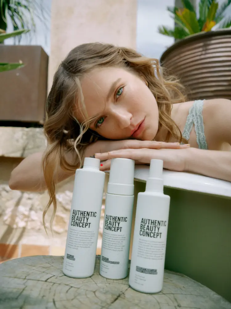 Modelo apoyada junto a productos de la gama Amplify de Authentic Beauty Concept en un entorno natural, mostrando una rutina de cuidado capilar para cabello fino.