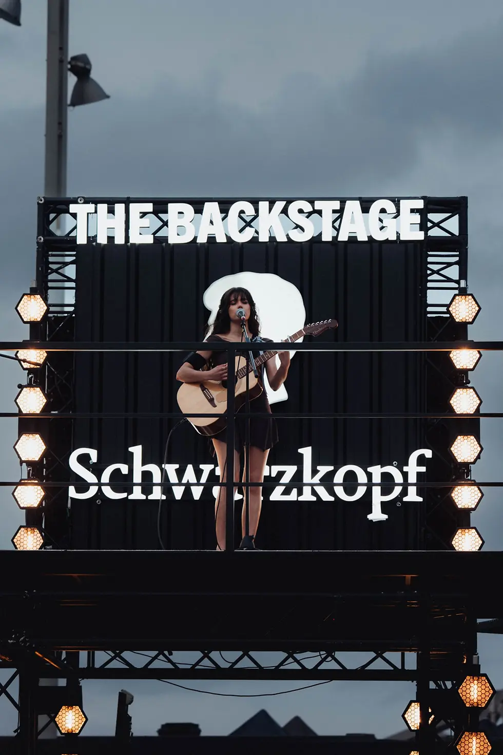 Escenario elevado con el rótulo “THE BACKSTAGE” y logotipo de Schwarzkopf Professional, donde una artista actúa con guitarra y micrófono durante el Primavera Sound Barcelona.