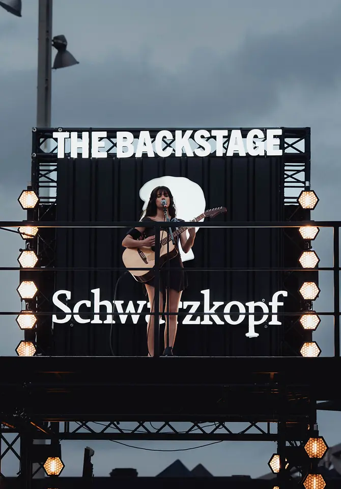 Escenario elevado con el rótulo “THE BACKSTAGE” y logotipo de Schwarzkopf Professional, donde una artista actúa con guitarra y micrófono durante el Primavera Sound Barcelona.