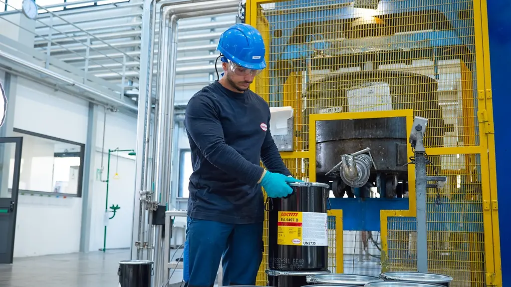 Interior industrial con operario manipulando y posicionando un recipiente de adhesivo LOCTITE EA 9497 junto a maquinaria en entorno de producción