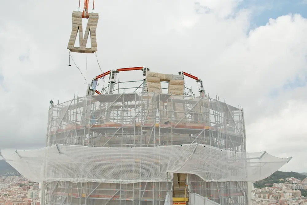 Proceso de instalación de un panel en la parte superior de la torre de Jesucristo de la Sagrada Familia con estructura en construcción y grúa elevando el elemento.