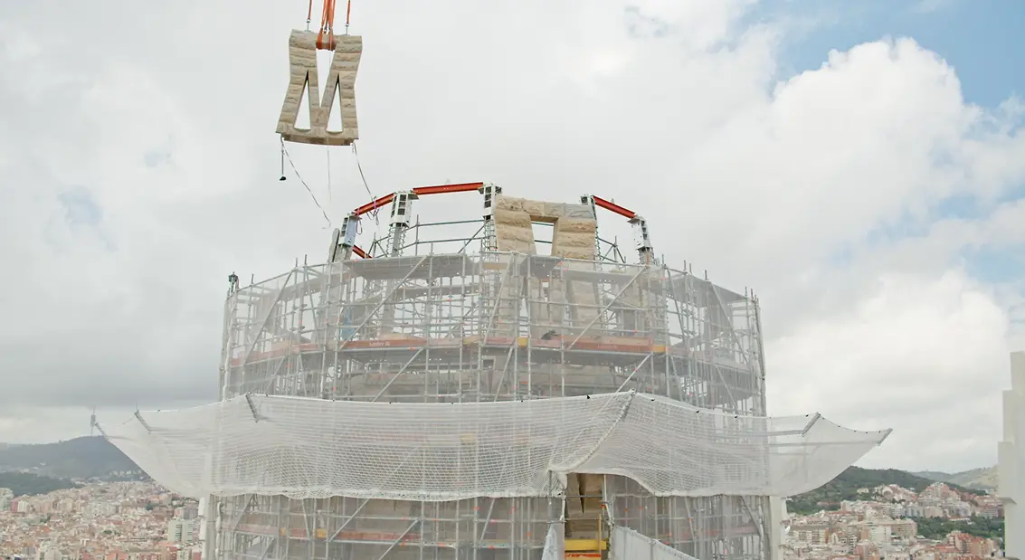Proceso de instalación de un panel en la parte superior de la torre de Jesucristo de la Sagrada Familia con estructura en construcción y grúa elevando el elemento.