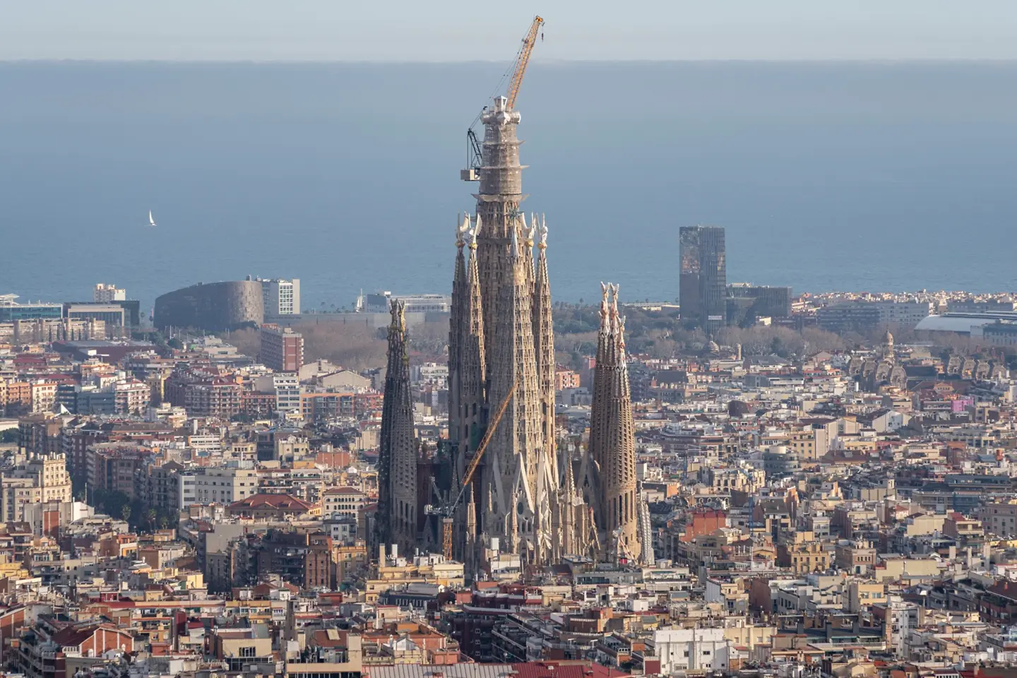 Vista general de la Sagrada Familia en Barcelona con sus torres centrales en construcción y cuatro personas posando en primer plano frente al templo en una plaza urbana con peatones y elementos de mobiliario urbano.
